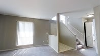 This two-story residence offers a functional and straightforward layout designed for comfortable everyday living. The main level features a spacious living area finished with soft wall-to-wall carpeting and neutral gray-toned walls that create a calm, understated ambiance. A large window fitted with vertical blinds allows natural light to pour into the room, brightening the space throughout the day. Adjacent to the living area, a tiled entryway leads to a front door, providing a clear and practical transition point between the interior and exterior of the home. A white-railed staircase rises from the main level to the second floor, with a skylight or upper window above the stairwell drawing additional natural light down into the lower level. The open sightlines between the living area, entry, and staircase give the main floor a connected, airy feel. The clean white trim and consistent neutral palette throughout contribute to a cohesive and move-in ready aesthetic. The second floor, accessible via the staircase, expands the overall footprint of the home, providing additional living or sleeping space above. Overall, the residence presents a clean, well-maintained interior with a practical two-level design suited for a range of lifestyles.