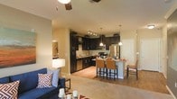 This single-floor residence showcases an open-concept layout that integrates the living, dining, and kitchen areas into one cohesive, functional space. The kitchen is a standout feature, outfitted with rich, dark espresso cabinetry that extends to the upper walls, stainless steel appliances including a refrigerator, microwave, and dishwasher, and under-cabinet track lighting that highlights the countertops below. A central kitchen island with bar-height seating provides additional prep space and serves as an informal dining area, accommodating several cushioned bar stools. Pendant lighting hangs above the island, adding a warm, focused glow to the space. The kitchen transitions naturally into a casual dining nook and a carpeted living area, where a ceiling fan overhead aids in air circulation. Light-toned walls keep the space feeling open and airy, while a mix of hard flooring in the kitchen and soft carpet in the living area delineates each zone. The overall ambiance is warm and inviting, blending modern finishes with a comfortable, functional design suited for everyday living.