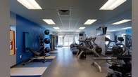 This single-floor fitness center offers a comprehensive workout environment with a practical, purpose-driven layout. The space is defined by its bold blue accent wall, which adds energy and visual character to the room, complemented by neutral gray flooring that extends throughout. A blue and white striped mat area near the free weights section provides a designated zone for floor exercises and stretching.

The cardio area lines one side of the room and features a row of treadmills and elliptical machines, all positioned to face wall-mounted televisions, making extended sessions more engaging. On the opposite side, a well-organized free weight and functional training area includes a rack of medicine balls and stability balls of various sizes, a weight bench, and dumbbells, offering versatility for strength and conditioning workouts.

Natural light filters in through glass doors at the far end of the room, providing a bright and welcoming atmosphere. The drop ceiling with evenly spaced recessed fluorescent panels ensures consistent overhead lighting throughout. A cable machine is also visible, rounding out the strength training options available in this fully functional fitness facility.
