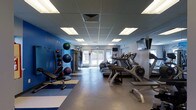 This single-floor fitness center offers a comprehensive workout environment with a practical, purpose-driven layout. The space is defined by its bold blue accent wall, which adds energy and visual character to the room, complemented by neutral gray flooring that extends throughout. A blue and white striped mat area near the free weights section provides a designated zone for floor exercises and stretching.

The cardio area lines one side of the room and features a row of treadmills and elliptical machines, all positioned to face wall-mounted televisions, making extended sessions more engaging. On the opposite side, a well-organized free weight and functional training area includes a rack of medicine balls and stability balls of various sizes, a weight bench, and dumbbells, offering versatility for strength and conditioning workouts.

Natural light filters in through glass doors at the far end of the room, providing a bright and welcoming atmosphere. The drop ceiling with evenly spaced recessed fluorescent panels ensures consistent overhead lighting throughout. A cable machine is also visible, rounding out the strength training options available in this fully functional fitness facility.