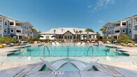 Located at 201 Sawyer Circle in Charleston, SC, this outdoor amenity space delivers a resort-style experience designed for relaxation and community gathering. The centerpiece is a generously sized swimming pool with crystal-clear blue water, complete with in-pool lounge chairs and entry steps, offering a leisurely retreat under the South Carolina sun.

Surrounding the pool, an expansive concrete sundeck is outfitted with modern sling-style chaise lounges and vibrant yellow accent stools, providing ample space for sunbathing and socializing. Navy blue market umbrellas are positioned throughout the deck, offering shaded seating areas alongside comfortable outdoor furniture groupings.

A standout feature of the space is the covered open-air pavilion, characterized by a vaulted wood-paneled ceiling, exposed timber beams, brick knee walls, and wall-mounted lighting. The pavilion houses a large flat-screen television and multiple seating arrangements, making it an ideal venue for outdoor entertainment.

Mature palm trees and carefully maintained landscaping frame the entire area, contributing to a tropical, welcoming ambiance. The surrounding multi-story residential buildings, finished in a classic coastal blue-grey palette with balconies and white trim, create an attractive and cohesive backdrop. The space benefits from abundant natural light and open sky views, reinforcing its resort-inspired character.