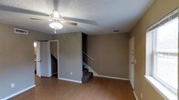 This two-floor residence offers a straightforward and functional layout spread across two connected levels. The main living area on the ground floor is finished with warm hardwood-style flooring that extends throughout the space, complementing the neutral tan-toned walls and white trim. A ceiling fan with integrated lighting fixture is centrally mounted on the textured popcorn ceiling, providing both airflow and ambient light to the room. A carpeted staircase with a handrail rises from the main living area, leading to the upper floor and serving as a central architectural element of the space. Doorways on the main level provide access to additional rooms, suggesting a closet or storage area alongside other living spaces. The entry door, visible to one side, features standard hardware and contributes to the overall practical design of the home. The overall ambiance is clean and understated, with a neutral color palette throughout that creates a versatile backdrop suitable for a variety of interior styles. The two-floor configuration allows for a natural separation between living and private spaces, making for an organized and livable environment.