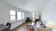 This single-floor space presents a clean, minimalist design centered around a well-lit living area. Large horizontal windows fitted with venetian blinds line one wall, allowing generous natural light to fill the room and creating an airy, open atmosphere. The flooring throughout is warm-toned hardwood, adding a sense of warmth that contrasts with the crisp white walls and ceiling.

The living area is thoughtfully arranged with a light-upholstered sofa dressed with patterned accent pillows, positioned across from a dark-framed television set on a low media console. A simple rectangular coffee table in a dark finish anchors the seating arrangement. A leather accent chair with clean lines adds a touch of contrast and additional seating to the space.

Decorative elements are kept minimal, including a tall branching plant arrangement and a potted green plant near the media area, contributing subtle organic texture to the otherwise neutral palette. A wall-mounted air conditioning unit is visible beneath the windows, alongside a baseboard heating system.

The overall ambiance is tidy, functional, and neutrally decorated, making it a versatile and comfortable living environment suited to a range of personal styles.