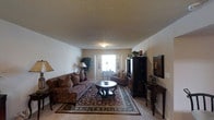 This single-story home offers a comfortable and classically styled interior designed for everyday living. The main living area serves as the heart of the home, showcasing a warm, neutral color palette with beige walls and a textured ceiling that adds subtle visual interest. Plush, patterned sofas are arranged around an ornate circular coffee table set atop a rich, decorative area rug, creating an inviting and cohesive gathering space. A tall, dark wood armoire adds a stately focal point to one side of the room, complemented by framed wall art and tasteful accent pieces that lend a traditional, curated feel. A slender console table along the wall provides additional display space for decorative items. Natural light filters through the entryway at the far end of the living room, where a front door flanked by sidelights creates a bright and welcoming threshold. The carpeted flooring runs throughout, contributing to the overall warmth and comfort of the space. Thoughtfully placed floor and table lamps ensure the room is well-lit during all hours. The overall ambiance is one of classic, understated elegance, making this a functional and inviting single-floor home.