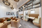 This elegantly designed single-floor communal space offers a refined yet inviting atmosphere across a generously proportioned open floor plan. At the heart of the space stands a dramatic floor-to-ceiling marble fireplace, flanked by plush tufted leather seating and anchored by a woven area rug beneath striking globe-shaped metallic pendant chandeliers, creating an intimate lounge area. Soaring ceilings amplify the sense of openness throughout.

Adjacent to the lounge, a series of built-in booth seating nooks with light wood paneling and sage-green upholstery are separated by architectural grid-paneled partitions, offering semi-private gathering spaces ideal for meetings or casual socializing. A dedicated dining area features a modern wood-top table with minimalist chairs, positioned near large windows that draw in abundant natural light and views of the surrounding greenery.

The reception area is highlighted by a sculptural wood-and-stone desk, vertical slatted wood columns, and a curated gallery wall arrangement. A bold teal accent wall adorned with organic wood-slice wall art adds a natural, artistic focal point. Large-format slate-toned tile flooring flows seamlessly throughout, unifying the space. The overall ambiance is contemporary and welcoming, balancing professional functionality with warm, residential-inspired design elements.