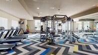 This fitness center is organized across two connected zones, each designed to support a full range of exercise routines. The main area is immediately distinguished by its striking chevron-patterned carpet in shades of gray, white, blue, and yellow, lending a dynamic and modern aesthetic to the space. Recessed lighting throughout keeps the environment bright and energized.

The primary workout zone houses a robust selection of Life Fitness strength equipment, including a large multi-station cable and weight machine system at its center. Flanking this are rows of cardio machines — treadmills, ellipticals, stationary bikes, and stair climbers — positioned near large windows that overlook outdoor greenery, allowing natural light to fill the space. Wall-mounted televisions are positioned near the cardio equipment for convenience. A dumbbell rack and free weights are also available near the entrance.

The secondary workout room features rich hardwood-style flooring and a quieter atmosphere suited for focused strength training. It includes additional cable machines and benches, along with a sauna room enclosed by warm wood-framed glass doors. Large windows with blinds line the walls, providing ample daylight while maintaining privacy. A glass entry door connects this room directly to an outdoor area.