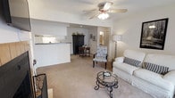 This single-floor residence offers a warm and functional living environment with an open-concept layout that seamlessly connects the main living spaces. The living area is generously sized, featuring soft wall-to-wall carpeting that adds comfort underfoot and a ceiling fan with integrated lighting that promotes air circulation and provides ambient illumination. A round glass-top coffee table with decorative ironwork anchors the seating arrangement, complemented by a classic white sofa adorned with accent pillows.

The space flows naturally into a dining or sitting area, where a traditional dark wood armoire and an antique-style side table with matching chair add a touch of classic elegance. The open pass-through kitchen features white cabinetry and a raised countertop bar, providing both a practical workspace and a visual boundary between the kitchen and the living area without sacrificing the open feel of the space.

Neutral, light-toned walls throughout the residence create a bright and airy atmosphere, while the understated color palette allows for easy personalization. A hallway visible in the background suggests access to additional private rooms, completing a well-organized and cohesive floor plan that balances openness with defined living zones.
