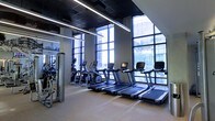 This contemporary fitness center offers a sleek, professionally designed workout environment that combines functionality with modern aesthetics. The space is anchored by a row of treadmills and elliptical trainers positioned along expansive floor-to-ceiling windows, which flood the interior with natural light and frame striking views of the surrounding urban cityscape. The large glass window wall creates an open, airy atmosphere that enhances the sense of spaciousness throughout the facility. Overhead, linear LED lighting is suspended from a dark ceiling grid, providing consistent, bright illumination across the workout floor. The light-toned flooring complements the industrial-modern design scheme, contrasting elegantly with the darker ceiling elements. Strength and resistance training equipment is arranged along the mirrored walls, which further amplify the sense of space and allow users to monitor their form during workouts. The mirrors extend across a full wall, reflecting both the equipment and the natural light from the windows. The overall ambiance is polished and professional, with a clean layout that ensures ease of movement between stations. This fitness center is thoughtfully designed to deliver an energizing and motivating workout experience within a high-end, urban setting.