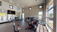 This single-story fitness center is designed to accommodate a comprehensive workout experience within a generously proportioned, open layout. The space is characterized by dramatically high ceilings that create an airy, energizing atmosphere, complemented by clerestory windows positioned near the roofline that draw in natural light from above. A striking exposed brick wall in a light, neutral tone serves as a prominent architectural feature, adding texture and visual warmth to the interior.

The main workout area is finished with durable rubberized flooring throughout, providing both comfort and safety during exercise. Spin bikes are arranged to take advantage of large, floor-to-ceiling glass panels and a roll-up garage-style door that opens toward outdoor greenery, flooding the space with natural light and views of the surrounding landscape. A wall-mounted ballet barre runs along the brick wall, accompanied by storage hooks for mats and accessories, as well as a nearby dumbbell rack.

Full-length mirrors along one wall visually expand the space and support strength training activities. Multiple access points include glass-paneled double doors and solid black interior doors, connecting to additional fitness areas and exterior walkways. Recessed lighting supplements the abundant natural light, ensuring a well-lit environment at all times. The overall ambiance is clean, modern, and functional.