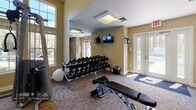 Located at 4133 Arbor Dr in Shrewsbury, MA, this single-story community facility is thoughtfully designed to accommodate a variety of recreational, fitness, and professional needs. The space is organized into several distinct functional areas, each offering a well-appointed environment for its intended use.

The centerpiece of the facility is a full indoor basketball court, distinguished by its soaring ceiling height, smooth tan sport-surface flooring with crisp white court markings, and regulation backboards. The walls feature a clean two-tone design in soft teal and white, complemented by elevated clerestory windows that introduce natural light and recessed fluorescent fixtures for consistent illumination throughout.

The fitness center is divided into two well-equipped zones: one housing strength-training machines, cable equipment, and free weights including a full dumbbell rack with mirrored walls and abundant natural light from large windows; the other featuring cardio machines such as ellipticals and steppers against bold teal and yellow accent walls.

The meeting and lounge area provides a professional setting with a large conference table, ergonomic seating, a wall-mounted television, a decorative fireplace, warm gold-toned walls, and built-in cabinetry with a wet bar. Carpeted corridors with warm neutral tones connect the various areas, completing this versatile, amenity-rich facility.