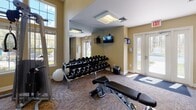 Located at 4133 Arbor Dr in Shrewsbury, MA, this single-story community facility is thoughtfully designed to accommodate a variety of recreational, fitness, and professional needs. The space is organized into several distinct functional areas, each offering a well-appointed environment for its intended use.

The centerpiece of the facility is a full indoor basketball court, distinguished by its soaring ceiling height, smooth tan sport-surface flooring with crisp white court markings, and regulation backboards. The walls feature a clean two-tone design in soft teal and white, complemented by elevated clerestory windows that introduce natural light and recessed fluorescent fixtures for consistent illumination throughout.

The fitness center is divided into two well-equipped zones: one housing strength-training machines, cable equipment, and free weights including a full dumbbell rack with mirrored walls and abundant natural light from large windows; the other featuring cardio machines such as ellipticals and steppers against bold teal and yellow accent walls.

The meeting and lounge area provides a professional setting with a large conference table, ergonomic seating, a wall-mounted television, a decorative fireplace, warm gold-toned walls, and built-in cabinetry with a wet bar. Carpeted corridors with warm neutral tones connect the various areas, completing this versatile, amenity-rich facility.