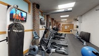 This fitness center offers a comprehensive workout environment housed within a distinctively designed space. Exposed brick columns punctuate the interior, complementing wood-trimmed windows that allow natural light to filter in alongside overhead fluorescent and skylight panels, keeping the space bright and energizing throughout.

The cardio zone is a standout feature, lined with a generous row of treadmills, elliptical trainers, and stationary bikes — many equipped with individual monitors for entertainment during workouts. Wall-mounted flat-screen televisions are positioned throughout for additional viewing convenience. A rowing machine and stair-climbing equipment further diversify the cardio offerings.

The strength training area includes a multi-station cable and weight machine system, a dedicated free weight section with a full dumbbell rack, a weight bench, and stability balls for functional training exercises. The flooring transitions between rubberized mat surfaces in workout zones and hard flooring near the entrance and locker area, providing both safety and practicality.

Toward the rear of the facility, wood-paneled locker room access and informational boards provide practical amenities for users. A ceiling-mounted fan and visible ductwork contribute to the space's ventilation. The overall ambiance blends industrial character with functional fitness design, creating an inviting and well-organized environment suitable for a full range of workout routines.