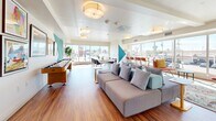 Located at 521 W Colorado St in Glendale, CA, this single-floor community lounge is a thoughtfully designed, multifunctional gathering space that combines modern aesthetics with practical amenities. Rich hardwood flooring runs throughout, providing a warm foundation that contrasts beautifully with the crisp white ceilings and walls.

The space is immediately distinguished by its bold, geometric wall murals rendered in teal, cobalt blue, red, and yellow, adding a dynamic and artistic energy to the interior. Flush drum ceiling fixtures provide consistent, even lighting that complements the generous natural light pouring in through large windows along the front-facing wall, offering open views of the surrounding Glendale streetscape.

The lounge is organized into several distinct zones. A comfortable seating area features a large sectional sofa with coordinating accent pillows, ideal for casual gatherings. A pair of high-back upholstered privacy chairs with side tables offers a quieter nook for focused work or conversation. A shuffleboard table and a wall-mounted TV add recreational and entertainment value. A dining and collaborative work area with tables and chairs supports group activities, while a built-in credenza with a wall-mounted display screen serves as a central information hub. A curated gallery wall of framed artwork further enhances the creative atmosphere of this well-appointed communal space.