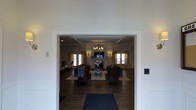 This single-floor space presents a sophisticated and well-appointed lounge or gathering room, visible through a wide, white-trimmed entryway that frames the interior with clean architectural precision. The room features a coffered ceiling with crisp white molding, adding a sense of structure and refinement overhead. A classic chandelier with warm-toned bulbs serves as a central focal point, complementing wall sconces that cast a soft, ambient glow throughout the space.

Dark hardwood flooring runs the length of the room, providing a rich contrast against the light walls and cream-toned drapery that frames multiple windows, allowing natural light to filter in from multiple sides. A traditional fireplace with a white mantel anchors the far wall, with a mounted flat-screen television above it, blending classic design with modern functionality.

The seating arrangement consists of leather club chairs and upholstered seating grouped around a central area rug, creating an intimate and inviting conversational layout. The overall ambiance is one of understated elegance, suited for relaxed gatherings or quiet retreats, with thoughtful design details throughout that reflect a commitment to quality and comfort.