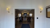 This single-floor space presents a sophisticated and well-appointed lounge or gathering room, visible through a wide, white-trimmed entryway that frames the interior with clean architectural precision. The room features a coffered ceiling with crisp white molding, adding a sense of structure and refinement overhead. A classic chandelier with warm-toned bulbs serves as a central focal point, complementing wall sconces that cast a soft, ambient glow throughout the space.

Dark hardwood flooring runs the length of the room, providing a rich contrast against the light walls and cream-toned drapery that frames multiple windows, allowing natural light to filter in from multiple sides. A traditional fireplace with a white mantel anchors the far wall, with a mounted flat-screen television above it, blending classic design with modern functionality.

The seating arrangement consists of leather club chairs and upholstered seating grouped around a central area rug, creating an intimate and inviting conversational layout. The overall ambiance is one of understated elegance, suited for relaxed gatherings or quiet retreats, with thoughtful design details throughout that reflect a commitment to quality and comfort.