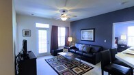 This single-story space centers around a well-appointed living room that immediately draws attention with its striking navy blue accent wall, which serves as a dramatic backdrop for the room's furnishings. A sleek black leather sofa and matching seating are arranged around a contemporary rectangular coffee table, anchoring the space with a modern aesthetic. A colorful geometric area rug adds visual interest and warmth to the carpeted floor beneath.

Natural light filters into the room through a large window and a glass-paneled door fitted with white blinds, both dressed with deep burgundy curtains that complement the bold wall color. A floor lamp positioned in the corner provides additional ambient lighting, while a ceiling fan with an integrated light fixture overhead ensures comfort and practical illumination throughout the space.

The walls are painted in a crisp, neutral tone, balancing the boldness of the accent wall and keeping the overall atmosphere bright and open. A framed piece of wall art adds a finishing touch to the navy feature wall, contributing to the room's polished, intentional design. The space conveys a comfortable and contemporary feel throughout.