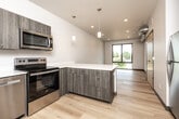 This thoughtfully designed single-floor unit offers a clean, modern aesthetic throughout its four rooms. Light hardwood flooring runs continuously across the entire space, complemented by crisp white walls and dark-trimmed doorways that add a contemporary contrast.

The open-concept kitchen serves as a functional centerpiece, equipped with a full suite of stainless steel appliances including an electric range, over-the-range microwave, dishwasher, and refrigerator. Wood-toned lower and upper cabinetry pairs with white quartz countertops, and a peninsula with pendant lighting above creates a natural division between the kitchen and living areas. Recessed lighting throughout ensures consistent, bright illumination.

The living and sleeping areas are well-proportioned and feature large, floor-to-ceiling black-framed windows that flood the rooms with natural light and offer views of the surrounding neighborhood. Exposed ductwork in one room adds an industrial touch to the otherwise minimalist design.

The bathroom is finished with the same light hardwood flooring and includes a walk-in shower with a curtain rod, toilet, and vanity. An in-unit washer and dryer is also present, adding practical convenience.

Overall, this space presents a cohesive modern design with quality finishes, ample natural light, and a functional layout.