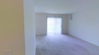 This single-floor living space features a clean, open layout with neutral white walls and wall-to-wall carpeting in a light beige tone, creating a simple and functional environment. The room is bathed in natural light, primarily sourced from a large sliding glass door fitted with white vertical blinds, which also provides direct access to an outdoor area. A smaller window positioned higher on the adjacent wall adds supplementary light and ventilation. The space is defined by smooth, plain walls and a structural column near the entry, subtly dividing the area without enclosing it. A floor-level air vent is visible along the baseboard near the sliding door, indicating a standard HVAC system integrated into the unit. The overall ambiance is minimal and neutral, offering a blank canvas well-suited for a variety of furnishing styles. The absence of decorative elements keeps the focus on the functional aspects of the space, including its open floor plan and access to natural light.
