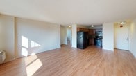 This single-floor residence offers a clean, functional layout with a cohesive design throughout. Light hardwood-style flooring runs continuously across all rooms, lending warmth and a sense of visual continuity to the space.

The open-concept living area is generously proportioned, with bright white walls and ample natural light streaming in. It flows seamlessly toward the kitchen, which is well-appointed with warm wood cabinetry, dark granite countertops, a striking mosaic tile backsplash, and a full suite of black appliances including a refrigerator, range, dishwasher, and built-in microwave hood. A flush-mount ceiling fixture provides overhead illumination.

The bedrooms are comfortably sized and filled with natural light from large windows dressed with vertical blinds. Each room features a wall-unit air conditioner, offering individual climate control. One bedroom includes a closet with sliding doors, while city views visible through the windows add an urban character to the living experience.

A hallway connects the rooms efficiently and features built-in upper cabinetry with modern hardware, providing additional storage. The corridor leads to the bright bedrooms, enhancing the sense of flow and accessibility throughout the unit. Overall, the space presents a practical yet comfortable urban living environment with thoughtful details and a neutral, versatile aesthetic.