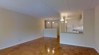 This single-floor residence presents a clean and functional layout with a warm, understated aesthetic. The living area is spacious and open, anchored by richly toned parquet flooring that adds classic character to the space. The neutral cream-colored walls and white ceilings keep the interiors feeling bright and airy, while subtle architectural framing defines the transition between living and kitchen zones.

A half-wall partition elegantly separates the living area from the adjacent kitchen, creating an open yet organized flow between the two spaces. The kitchen features a countertop bar ledge, white appliances including a refrigerator, and overhead pendant lighting that adds a warm, ambient glow. A wall-mounted sconce near the kitchen entrance further enhances the inviting atmosphere.

Pendant lighting also hangs in the background near what appears to be a hallway or entryway, hinting at additional rooms beyond the main living area. The polished parquet floors throughout reflect the interior lighting, lending the space a sense of warmth and depth. Overall, this residence combines practical design with timeless finishes to create a comfortable and cohesive living environment.