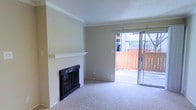 This single-floor space presents a clean, well-maintained interior with a straightforward layout designed for comfortable everyday living. The main living area is anchored by a wood-burning fireplace framed by a classic white mantel and contrasting black surround, adding a focal point and a warm, inviting atmosphere to the room. Crown molding runs along the upper perimeter of the walls, lending a subtle architectural detail that elevates the overall finish of the space.

Natural light fills the room through a full-width sliding glass door, which opens to an outdoor balcony enclosed by a wood-toned railing. The view outward provides a sense of openness and a connection to the surrounding environment. The sliding door also allows for convenient indoor-outdoor flow, making the balcony a practical extension of the living area.

Underfoot, plush wall-to-wall carpeting in a neutral beige tone adds softness and warmth throughout. The walls are finished in a light, neutral palette, complementing the textured ceiling and creating a bright, airy feel. The overall ambiance is calm and move-in ready, offering a versatile canvas adaptable to a variety of furnishing styles.