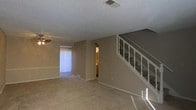 This two-story space features a carpeted main living area with neutral beige walls and a textured popcorn ceiling. A white-railed staircase with decorative spindles leads to the upper floor, serving as a functional and visually notable architectural element within the room. A ceiling fan is installed overhead, contributing to airflow and comfort throughout the space.

Natural light enters through a window dressed with vertical blinds, brightening the living area. An arched doorway connects the main living space to an adjacent room, adding a subtle architectural detail that breaks the straight lines of the interior. Recessed vents are positioned throughout the ceiling and floors, indicating a central HVAC system. Baseboards in white trim the perimeter of the room, providing a clean contrast to the beige walls and carpeted flooring.

The overall ambiance is practical and straightforward, with a layout that balances open living space on the main floor with additional living quarters accessible via the staircase on the second floor.