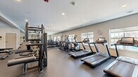 Located at 3701 Lilac Ln in Enola, PA, this fitness center offers a comprehensive workout facility across a single, open floor. The space is designed to accommodate a wide range of exercise needs, with a thoughtful layout that separates cardio and strength training zones. The cardio area features a row of treadmills and elliptical machines, each equipped with individual display consoles, positioned along windows that allow natural light to fill the room and offer outdoor views. A wall clock keeps members on schedule during their workouts. The strength training zone is outfitted with Matrix cable machines, a power rack with Olympic barbell and weight plates, adjustable benches, and a full dumbbell rack with a comprehensive range of free weights. Stability balls, resistance tools, and accessories are neatly organized on wall-mounted storage racks, supporting functional and flexibility training. Full-length mirrors line one wall, enhancing the sense of openness while allowing users to monitor their form. The flooring throughout is durable, dark rubberized material suited for heavy equipment use. Recessed lighting provides consistent, even illumination across the space. The overall ambiance is clean, functional, and professional, making this fitness center a well-rounded facility for residents seeking a complete exercise experience.