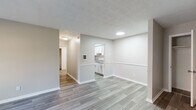 This 1-bedroom, 1-bathroom unit in Lexington, Kentucky, is laid out on a single floor with a clean, modern aesthetic and a neutral color palette throughout. The living area is open and well-lit, featuring wood-look luxury vinyl plank flooring, white trim, and a textured ceiling. A wall-mounted A/C unit and a large window with blinds allow natural light to fill the room. The layout connects the living space to a defined dining area, separated by a half-wall that opens directly to the kitchen. The kitchen is equipped with white shaker-style cabinetry, granite-patterned countertops, a black electric range, a full-size refrigerator, and an overhead microwave, all set atop matching wood-look tile flooring. A hallway leads to the bedroom, which features soft carpeting, a ceiling fan, and a window with a natural view. The full bathroom is finished with wood-look tile flooring, a white vanity with a sink and mirror, and a separate toilet area. A closet is accessible off the bathroom hallway, providing additional storage. The unit presents a well-maintained, move-in-ready interior with consistent finishes and a functional, efficiently designed floor plan.