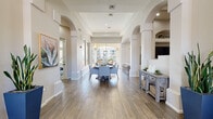 This single-floor community clubhouse is designed with an open, welcoming layout that seamlessly connects multiple functional areas. The main corridor features elegant arched doorways and structural columns that frame a spacious dining area at its center, complete with round tables and upholstered chairs. Large windows and a glass-enclosed view toward an outdoor courtyard flood the space with natural light, creating an airy atmosphere throughout.

The lounge areas offer comfortable seating arrangements, including plush sofas and accent chairs, complemented by botanical artwork and warm, neutral tones. A standout feature is the tiled fireplace set within an arched alcove, paired with a wall-mounted television — ideal for relaxed gatherings. Adjacent to this is a well-equipped kitchenette with white shaker cabinetry, granite-style countertops, a full sink, compact appliances, and rustic floating shelves for display.

Wood-look tile flooring runs consistently throughout, lending warmth and cohesion to every area. Built-in shelving niches add architectural character, while strategic recessed lighting ensures each zone is well-lit. A fitness center, visible through large glass windows, provides additional amenity space. A private office and multiple seating nooks round out this thoughtfully designed, multi-use community facility.