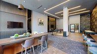 This contemporary amenity lounge is designed with a sharp, urban aesthetic that blends social gathering spaces with recreational entertainment. The single-floor layout is anchored by a striking entrance lobby featuring floor-to-ceiling glass windows that flood the space with natural light and offer street-facing views. A large-scale, vivid mural adorns one wall of the entry area, setting an artistic tone that carries throughout the space, complemented by additional curated artwork displayed on the walls.

The main lounge area is thoughtfully zoned, offering distinct areas for lounging, dining, and recreation. A seating nook provides a relaxed atmosphere with plush chairs arranged around a flat-screen television, framed by a dramatic slatted wood accent wall with integrated linear LED lighting. Nearby, a billiards table with a bold red felt surface serves as a central recreational feature. A bar-height counter with stylish upholstered stools offers additional seating, while high-top tables provide casual dining options.

Flooring throughout transitions between large-format stone-look tiles and warm wood tones, with a patterned area rug anchoring the central lounge zone. Recessed linear ceiling lights enhance the modern, polished ambiance, and glass-paneled interior doors connect to adjacent spaces, maintaining an open and airy flow throughout.