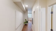 This well-designed single-floor unit offers a cohesive modern aesthetic characterized by clean lines, a neutral warm palette, and wood-look flooring that flows seamlessly throughout the space. The entry hallway sets an inviting tone, featuring recessed circular lighting, crisp white walls, and tasteful wall art that leads naturally into the main living areas. The kitchen is efficiently laid out and outfitted with light natural wood cabinetry, white countertops, and a full suite of stainless steel appliances including a refrigerator, electric range, over-the-range microwave, and dishwasher. A window above the counter brings in natural light, enhancing the kitchen's bright and functional feel. The bedroom is a comfortable, well-lit retreat with a large window that allows abundant natural light to fill the room. It comfortably accommodates a bed and a workspace, making it versatile for both rest and productivity. The bathroom is clean and modern, featuring a floating wood-toned vanity with an integrated white sink, a large wall mirror with overhead lighting, grab bars, and a shower-tub combination. Recessed lighting throughout the unit ensures consistent, even illumination, while the open layout between the kitchen and living area promotes an airy, connected sense of space.