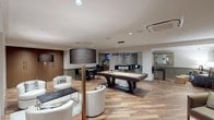 The Club Lounge at 1 St. Mary's Convent in Peekskill, NY is a thoughtfully designed amenity space that seamlessly blends elegance with functionality across several distinct zones. Wide-plank hardwood flooring in a warm, mixed-tone finish runs throughout, unifying the space and lending it a refined yet inviting character.

At the heart of the lounge is a central game area anchored by a full-size billiards table beneath a sleek rectangular pendant light. Adjacent to this is a wet bar and kitchenette, finished with richly textured stone-look wall cladding, a granite countertop, bar seating, stainless steel appliances, and a wall-mounted television — ideal for entertaining.

A comfortable lounge area features a generous sectional sofa, swivel accent chairs, and a glass-top coffee table, all set against a striking dimensional accent wall with a geometric raised-relief pattern. Curated artwork and brass floor lamps add warmth and visual interest throughout.

A separate dining area with a crisp white table and upholstered chairs opens directly into the main lounge through wide doorways, offering a more intimate setting. A foosball table and additional seating areas round out the recreational offerings. Recessed lighting throughout ensures a well-lit, polished atmosphere suited for both casual gatherings and organized events.