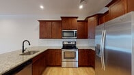 Located in Tewksbury, MA, this single-floor residence offers a thoughtfully designed layout with quality finishes throughout. The kitchen is a standout feature, showcasing rich cherry wood cabinetry, granite countertops, a white subway tile backsplash, and a center island with a built-in sink. Stainless steel appliances — including a range with over-the-range microwave and a French door refrigerator — complete the space. Light hardwood flooring flows through the kitchen and into the open living area, which is highlighted by crown molding, recessed lighting, a ceiling fan, and direct access to the outdoors through a glass-panel door. The bedrooms feature plush carpeting, ceiling fans, and windows that bring in natural light and neighborhood views. The bathrooms are well-appointed with granite-topped white vanities, tile flooring, and oil-rubbed bronze fixtures; the primary bath includes a walk-in shower with a bold black-framed glass door. A dedicated laundry room with side-by-side washer and dryer and wire shelving adds everyday convenience. The consistent use of neutral tones and warm wood accents gives the entire space a clean, cohesive, and inviting character.
