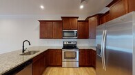 Located in Tewksbury, MA, this single-floor residence offers a thoughtfully designed layout with quality finishes throughout. The kitchen is a standout feature, showcasing rich cherry wood cabinetry, granite countertops, a white subway tile backsplash, and a center island with a built-in sink. Stainless steel appliances — including a range with over-the-range microwave and a French door refrigerator — complete the space. Light hardwood flooring flows through the kitchen and into the open living area, which is highlighted by crown molding, recessed lighting, a ceiling fan, and direct access to the outdoors through a glass-panel door. The bedrooms feature plush carpeting, ceiling fans, and windows that bring in natural light and neighborhood views. The bathrooms are well-appointed with granite-topped white vanities, tile flooring, and oil-rubbed bronze fixtures; the primary bath includes a walk-in shower with a bold black-framed glass door. A dedicated laundry room with side-by-side washer and dryer and wire shelving adds everyday convenience. The consistent use of neutral tones and warm wood accents gives the entire space a clean, cohesive, and inviting character.