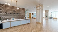 Located at 3955 Design Center Drive in Palm Beach Gardens, Florida, The Marc presents an expansive single-floor amenity space thoughtfully designed with a coastal-contemporary aesthetic. The space opens at a welcoming reception desk, framed by a striking textured feature wall bearing the property's branding and flanked by decorative vertical-slatted columns clad in warm wood tones. Intricate parquet-patterned flooring in light natural wood flows seamlessly throughout, tying each zone together cohesively.

The generous lounge area is anchored by a large sectional sofa and plaid accent chairs arranged atop a geometric area rug, all set beneath soaring ceilings and floor-to-ceiling windows draped with soft teal sheer curtains that invite abundant natural light and outdoor views. A game area nearby features a sleek white billiards table surrounded by lush tropical potted plants against another wall of expansive glazing.

A fully equipped catering kitchen showcases a bold blue subway tile backsplash, stainless steel appliances, a white waterfall island with teal counter seating, and warm wood cabinetry. A glass-enclosed private meeting or dining room with brick accent walls and pendant lighting provides an intimate, sophisticated retreat within the broader space. Greenery is thoughtfully distributed throughout, enhancing the bright, airy ambiance of the entire floor.