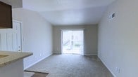 This single-floor space features a clean, straightforward layout centered around a comfortable living area. The room is finished with wall-to-wall carpet in a neutral tone, complementing the light beige walls and white baseboards to create a cohesive, understated aesthetic. A sliding glass door fitted with vertical blinds serves as the focal point of the main living area, allowing natural light to fill the room while providing direct access to an outdoor space, visible through the glass. The ceiling features a subtle angled design, adding a modest architectural detail to the otherwise simple interior. The space is move-in ready, with a neutral palette that offers flexibility for a variety of furnishing styles. Electrical outlets are conveniently positioned along the walls, and the overall layout feels open and uncluttered. The combination of natural light from the sliding door and the soft, neutral finishes throughout gives the space a calm, welcoming ambiance suitable for everyday living.