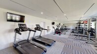This fitness center in Pikesville, MD offers a thoughtfully designed workout space across a single open floor. The room features a clean, modern aesthetic with crisp white walls, recessed lighting, and a patterned multi-tone carpet that adds visual interest while providing a comfortable exercise surface. A large wall-to-ceiling mirror spans one side of the room, creating a sense of expanded depth and allowing users to monitor their form during workouts.

The cardio zone is well-represented with multiple Spirit treadmills and a row of upright stationary bikes and elliptical trainers lined along the mirrored wall, each equipped with individual monitors. Strength training options include a well-stocked free weights area with a tiered rack holding kettlebells, medicine balls, and exercise balls in varying sizes, along with a multi-function cable and weight machine. A rowing machine and additional functional training equipment round out the facility's offerings.

A wall-mounted water fountain with a bottle-filling station is conveniently accessible within the space. Natural light filters in through a window on one end, complementing the recessed overhead lighting. A dedicated entrance door provides direct access, and the overall layout ensures ample open floor space for stretching or floor exercises, making this fitness center a comprehensive and functional facility.