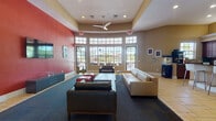 This well-appointed single-floor amenity space is designed to serve as a social and entertainment hub. The centerpiece is a generously sized lounge area distinguished by a dramatic red accent wall, a wall-mounted flat-screen television, and a large bank of floor-to-ceiling gridded windows that curve along one wall, flooding the room with natural light and providing views of an outdoor pool area. Comfortable leather seating arrangements are oriented around a central coffee table atop a dark area rug, while a ceiling fan overhead adds a relaxed, casual atmosphere. Tile flooring transitions seamlessly throughout the space.

Adjacent to the lounge is a fully equipped kitchen featuring rich wood cabinetry, granite countertops, a stainless steel refrigerator, microwave, and a popcorn machine—suggesting a connection to entertaining. A granite-topped breakfast bar with modern bar stools provides an informal seating option.

Casual dining nooks adorned with large coastal-themed photographic wall art and high-top tables offer additional gathering spots. A dedicated private screening room rounds out the entertainment offerings, equipped with tiered rows of black upholstered theater seats, acoustic wall panels, and a ceiling-mounted projector.