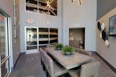 This contemporary communal space makes a strong architectural impression with its dramatic double-height ceilings, which lend an open, airy quality to the interior. Large glass-paneled doors along one wall introduce natural light and provide a visual connection to the exterior, enhancing the sense of openness throughout.

At the heart of the space sits a substantial concrete-top dining table surrounded by upholstered chairs in a neutral tone, creating a comfortable yet refined setting for group gatherings. Potted greenery placed along the table adds a touch of warmth and texture to the otherwise sleek, minimalist aesthetic.

A built-in wet bar area is tucked into an alcove along one wall, finished with a textured mosaic tile backsplash in earthy tones and equipped with under-counter refrigeration and cabinetry — making it well-suited for entertaining. The flooring features a patterned carpet in muted tones, grounding the space with subtle visual interest.

Overhead, an upper-level mezzanine or clerestory element is visible, fitted with industrial-style track lighting that contributes to the modern design language. The overall ambiance is polished and functional, with a clean, neutral palette throughout.