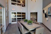 This contemporary communal space makes a strong architectural impression with its dramatic double-height ceilings, which lend an open, airy quality to the interior. Large glass-paneled doors along one wall introduce natural light and provide a visual connection to the exterior, enhancing the sense of openness throughout.

At the heart of the space sits a substantial concrete-top dining table surrounded by upholstered chairs in a neutral tone, creating a comfortable yet refined setting for group gatherings. Potted greenery placed along the table adds a touch of warmth and texture to the otherwise sleek, minimalist aesthetic.

A built-in wet bar area is tucked into an alcove along one wall, finished with a textured mosaic tile backsplash in earthy tones and equipped with under-counter refrigeration and cabinetry — making it well-suited for entertaining. The flooring features a patterned carpet in muted tones, grounding the space with subtle visual interest.

Overhead, an upper-level mezzanine or clerestory element is visible, fitted with industrial-style track lighting that contributes to the modern design language. The overall ambiance is polished and functional, with a clean, neutral palette throughout.