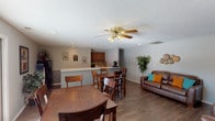 This single-story space in Georgetown South offers a well-organized, open-concept layout that seamlessly connects the living, dining, and kitchen areas. The main living area features hardwood-style flooring throughout, a textured ceiling, and recessed lighting complemented by a ceiling fan with a built-in light fixture. Brown leather seating, including a sofa and armchair, are arranged to create a comfortable gathering space, accented by colorful throw pillows and decorative wall art.

Two dining areas are present within the open floor plan — one positioned near French doors that allow natural light to pour in and provide views of the outdoors, and another situated adjacent to the kitchen peninsula. The peninsula serves as a natural divider between the kitchen and the main living space, offering counter seating and additional workspace.

The kitchen features warm wood cabinetry, a tile backsplash, black appliances including a gas range, and recessed lighting. A hallway visible from the living area leads to additional rooms and an exterior door. Greenery throughout the space adds a fresh, inviting touch, contributing to the overall warm and functional atmosphere of this single-floor layout.