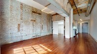 This single-floor loft masterfully combines raw industrial heritage with modern design sensibilities. The expansive main living area immediately commands attention with its soaring ceilings, exposed structural beams, and sweeping brick walls that tell the story of the building's storied past. Rich, warm-toned hardwood flooring runs throughout, reflecting the abundant natural light that pours in through a pair of tall arched windows framed in dark metal — a defining architectural feature that adds both grandeur and character. Recessed rectangular LED lighting overhead provides a clean, contemporary counterpoint to the space's historic textures. The kitchen is sleek and well-appointed, featuring white cabinetry with modern hardware, glass tile backsplash, stone countertops, and stainless steel appliances including a range, microwave, and dishwasher. A dedicated closet space is accessible through a sliding door, offering practical storage. The bathroom is finished to a high modern standard, with large-format gray tile flooring and walls, a walk-in shower with a glass sliding door, and a clean white vanity with modern fixtures. The overall ambiance throughout this loft is one of urban sophistication — raw and refined in equal measure.