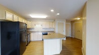 This single-story space showcases an open-concept kitchen designed for both functionality and comfort. The kitchen is outfitted with light maple cabinetry that lines the perimeter walls, offering generous storage throughout. Granite-style countertops provide ample workspace along the cabinetry run, while a centrally positioned kitchen island extends additional prep and seating space into the room. Black stainless appliances, including a refrigerator, gas range, and dishwasher, are integrated seamlessly into the layout. A double sink is situated beneath the cabinetry, accompanied by under-cabinet lighting that illuminates the workspace below. Recessed lighting and a flush ceiling fixture ensure the kitchen remains well-lit throughout. The warm-toned hardwood flooring flows continuously from the kitchen into the adjacent living areas, creating a cohesive and unified aesthetic. The open layout connects the kitchen to additional rooms visible in the background, with interior doors leading to separate spaces. The overall ambiance is clean, bright, and practical, with a neutral color palette that lends a welcoming and versatile atmosphere throughout the space.