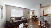 This single-floor residence offers a thoughtfully designed layout that balances comfort and functionality throughout. The open-concept living and dining area serves as the heart of the home, featuring warm hardwood-style flooring, neutral taupe walls, and large windows with white blinds that allow abundant natural light to fill the space. The living area provides a welcoming atmosphere, while the adjacent dining space accommodates a round table with seating for four. A peninsula-style breakfast bar connects the living and dining areas to the kitchen, which is equipped with white cabinetry, granite-style countertops, a stainless steel sink, and black appliances including a full-size refrigerator. The kitchen also features matching hardwood-style flooring and direct access to a hallway. A secondary sitting room offers plush carpeting, additional windows, and a relaxed ambiance ideal for a reading nook or home office. The bedroom is carpeted and includes a ceiling fan, a sliding glass door with vertical blinds, and ample space for a full suite of furniture. Two bathrooms serve the unit, each featuring white vanity cabinetry, granite-style countertops, and a shower with a curtain. The consistent use of neutral tones and natural-look flooring creates a cohesive and inviting atmosphere throughout the entire space.