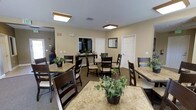 This single-floor clubhouse serves as a warm and inviting community gathering space, thoughtfully designed to encourage socializing and relaxation. The main area features multiple dining tables with granite-style surfaces, each accompanied by dark wood chairs with cushioned seats, arranged to comfortably accommodate groups of varying sizes. Lush potted greenery is placed throughout, adding a fresh, welcoming touch to the neutral-toned interior. The walls are finished in a soft beige palette, complemented by white trim and doors that lend a clean, classic aesthetic. Recessed flush-mount lighting fixtures provide consistent, even illumination across the space. A console table along the back wall offers a decorative accent, while a framed mirror adds depth to the room. Additional doorways suggest access to supplementary amenities or utility areas. The flooring features a neutral carpet that ties the room together and contributes to a quiet, comfortable atmosphere. Overall, the clubhouse presents a practical yet pleasant environment well-suited for community gatherings, casual dining, and everyday socializing.
