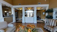 This single-floor space presents a sophisticated and welcoming atmosphere, centered around a beautifully appointed lobby or reception area. The space is defined by prominent white columns that frame a central reception desk, finished in crisp white cabinetry and positioned beneath an arched transom window that draws in natural light from the entrance. Warm golden-tan walls create an inviting ambiance throughout, complemented by flush-mount ceiling light fixtures set within coffered ceiling details. The flooring transitions between rich hardwood and dark tile accents, adding visual interest and depth to the open layout. Decorative framed artwork adorns the walls on either side of the reception area, lending a polished, curated feel to the space. A seating area is visible in the foreground, accessorized with a floral arrangement in warm amber tones atop a patterned area rug, enhancing the welcoming character of the environment. The open sightlines and well-defined zones within this lobby create a sense of spaciousness, making it well-suited for greeting visitors while maintaining an organized and professional aesthetic.