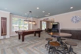 Located at 1010 Flats Ave in Mebane, NC, this single-story community clubhouse offers a well-rounded amenity space designed for both recreation and productivity. The layout is anchored by a spacious game room with a distinctive slate-blue accent ceiling, recessed lighting, and herringbone-patterned flooring that lends a polished, contemporary feel. This central gathering area features a full-size pool table beneath a classic billiard pendant light, a vintage arcade cabinet, high-top tables with industrial-style stools, and a wet bar area with dark cabinetry and a mini refrigerator — ideal for social gatherings. Natural light pours in through multiple windows fitted with white blinds, brightening the open interior. A separate dining room provides a more intimate setting, complete with a round dark-wood table, leather-upholstered chairs, crown molding, and a tasteful pendant light. The business center offers a built-in wooden desk with dual computer workstations, a printer, and a corkboard, catering to residents' professional needs. A water fountain station with a bottle-filling feature is also conveniently accessible. Throughout the space, neutral wall tones and crown molding details contribute to a clean, inviting ambiance.