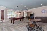 Located at 1010 Flats Ave in Mebane, NC, this single-story community clubhouse offers a well-rounded amenity space designed for both recreation and productivity. The layout is anchored by a spacious game room with a distinctive slate-blue accent ceiling, recessed lighting, and herringbone-patterned flooring that lends a polished, contemporary feel. This central gathering area features a full-size pool table beneath a classic billiard pendant light, a vintage arcade cabinet, high-top tables with industrial-style stools, and a wet bar area with dark cabinetry and a mini refrigerator — ideal for social gatherings. Natural light pours in through multiple windows fitted with white blinds, brightening the open interior. A separate dining room provides a more intimate setting, complete with a round dark-wood table, leather-upholstered chairs, crown molding, and a tasteful pendant light. The business center offers a built-in wooden desk with dual computer workstations, a printer, and a corkboard, catering to residents' professional needs. A water fountain station with a bottle-filling feature is also conveniently accessible. Throughout the space, neutral wall tones and crown molding details contribute to a clean, inviting ambiance.