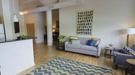 This single-floor space showcases a distinctive industrial loft aesthetic, highlighted by exposed cylindrical structural columns and white-painted ductwork running across the ceiling. The open-concept layout connects the living area to a visible bedroom in the background, creating a sense of spaciousness and continuity throughout.

The living area is anchored by light hardwood flooring, which adds warmth to the industrial character of the space. A bold chevron-patterned area rug in navy and cream defines the seating zone, where a contemporary light gray sofa is styled with green, yellow, and blue accent pillows. A round wooden coffee table with a lower shelf sits nearby, providing both functionality and visual interest.

Natural light fills the space, reflecting off the pale walls and light floors. A large abstract artwork featuring navy brushstroke shapes serves as a focal point on the main wall. The partially visible kitchen area to the left features dark countertops, offering a subtle contrast to the bright, airy surroundings.

The bedroom area, glimpsed through the open layout, features a low-profile bed and warm ambient lighting, maintaining the cohesive, relaxed atmosphere found throughout the space.