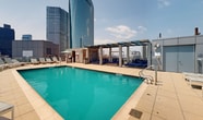 This expansive rooftop space is a testament to urban luxury and innovative design. Situated atop a modern high-rise, it offers a multi-faceted outdoor experience with breathtaking panoramic views of the city skyline. The centerpiece is a sleek, azure swimming pool flanked by comfortable loungers, perfect for relaxation or sunbathing. Multiple seating areas are thoughtfully arranged, featuring a mix of modern and rustic furniture, from plush sofas to woven chairs and wooden accents. A curved walking path lined with lush greenery adds a touch of nature to the urban setting. The space is further enhanced by string lights and modern lighting fixtures, creating an inviting ambiance for day and night use. Glass balustrades ensure safety without obstructing the spectacular views. Additional amenities include covered seating areas and what appears to be a bar or kitchen space, making this rooftop an ideal venue for both personal relaxation and social gatherings. The juxtaposition of the natural elements against the backdrop of towering skyscrapers creates a unique and captivating urban oasis.